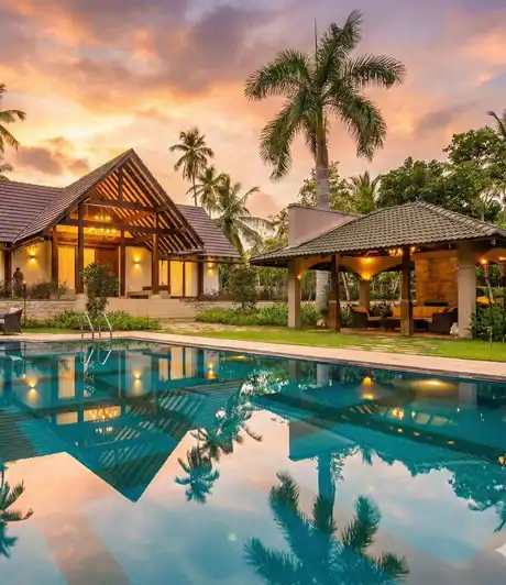Elegant villa with swimming pool surrounded by palm trees and lush greenery at sunset.